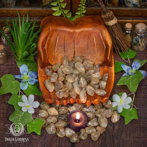 Many specimens of the "Scapolite Tumbled Gemstone" product are spilling from carved wooden cupped hands. They are surrounded by faux greenery and a tealight candle. They are tumbled smooth and are partially transparent in shades of brown and gold with dark inclusions. Presented by Inked Goddess Creations.