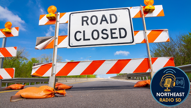 Road closed sign with orange barricades on a highway; blue sky and trees in the background; "Northeast Radio SD" logo in the corner.