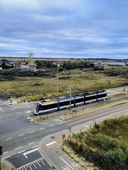 Das Bild zeigt den Blick aus dem Fenster in die andere Richtung: einen Aussichtsturm in einer Seegras-Düne davor eine geteilte Straße in deren Mitte die Küstenstraßenbahn gerade fährt
