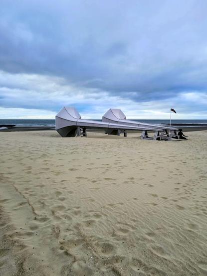 Auf dem Bild zu eehen ist das Kunstwerk" Ich kann es hören" im.Original " I Can Hear It" von  Ivars Drulle bestehend aus zwei nebeneinader liegenden übergroßen Hörtrichtern am Strand. Vor einem Trichter kniet eine Frau aus flachem schwarzem Blech und lauscht.