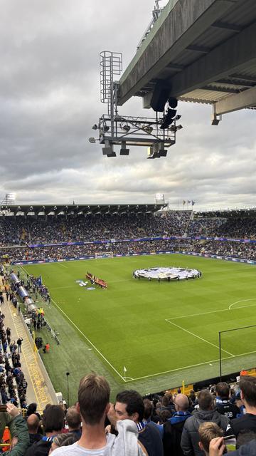 A packed stadium during a football match, with fans in the stands and teams lined up on the field beneath a cloudy sky. A large ceremonial banner is displayed at center field, with stadium lights on and a camera rig hanging overhead.