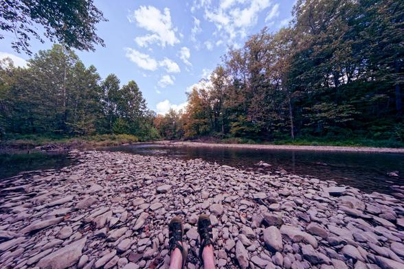Wide angle shot where a person's feet in Keen water shoes can be seen at the very bottom looking very tiny,  they're sitting on a field of angular rocks that is surrounded by brown-looking water on both sides,  the creek is bounded by trees on both sides that are starting to turn into fall color and above the sky is mostly blue with just scattered white clouds.