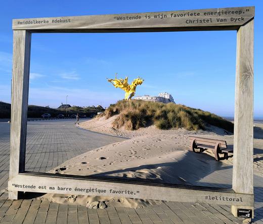 Das Bild zeigt das Kunstwerk "Lelittoral" von  Christel Van Dyck, das am Strand steht. Es besteht aus einem übergroßen Bilderrahmen, durch den man eine gelbe abstrakte Skulptur sieht die auf einer Seegrasdüne steht.