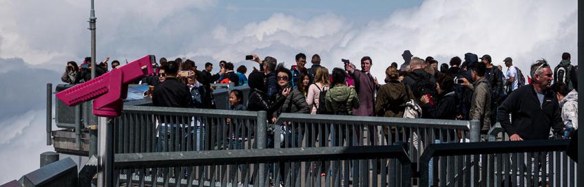 Viele Menschen stehen auf einer Aussichtsplattform, die von einem grauen Metallgeländer umgeben ist. Der Himmel im Hintergrund ist bewölkt und ein wolkenverhangener Bergkamm ist zu erkennen. Die meisten Menschen sind von hinten oder der Seite zu sehen. Eine Frau mit einer dunklen Sonnenbrille, die im vorderen Teil der Gruppe steht, blickt in die Kamera. Auf der linken Seite der Plattform steht ein grosser, pinkfarbener Binokular-Kasten auf einem silbernen Ständer. Fast alle Menschen tragen Freizeitkleidung wie Jacken und T-Shirts. Einige von ihnen machen Fotos mit ihren Handys.