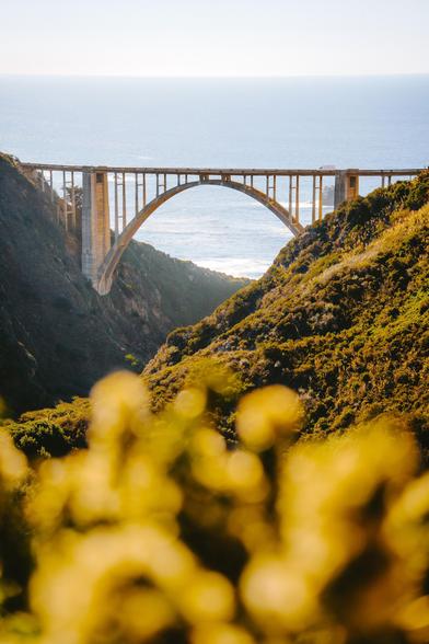 A large arch bridge spans a canyon with lush green hills on either side, overlooking the ocean in the distance.