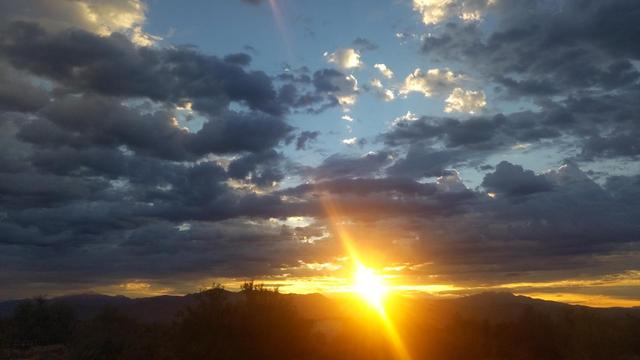 A dramatic sunrise over a mountainous desert landscape, with the sun low on the horizon casting bright golden rays through thick, dark clouds. The sky shifts from vivid yellow near the horizon to soft blue above, with cloud edges glowing silver and gold. Silhouetted hills and sparse shrubs frame the foreground.