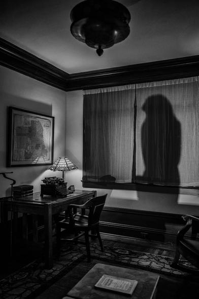 Darkened apartment with shadow of Maltese Falcon projected on window. The furnishings include a desk with typewriter, Tiffany style lamp, alarm clock, and above that, a map of San Francisco on the wall.