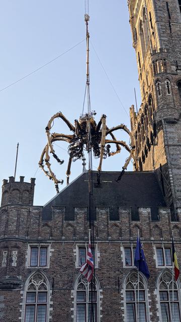 A large mechanical spider is being hoisted by a crane above a historic building. The structure features brick architecture with arched windows and multiple flags displayed. The sky is clear.