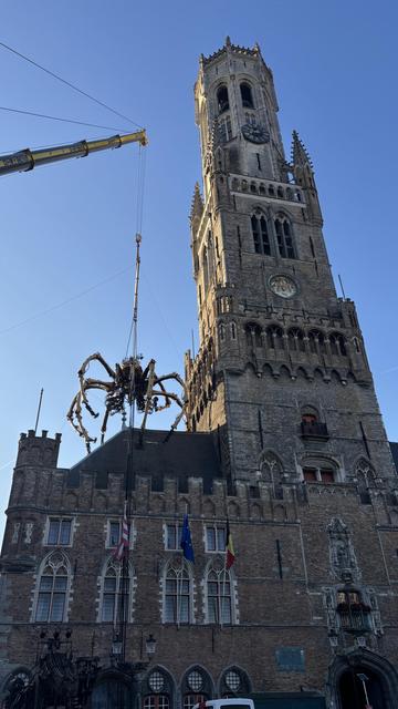 A large, mechanical spider is being lifted by a crane next to a tall, historic bell tower. The scene features a clear blue sky and includes several flags at the base of the tower.