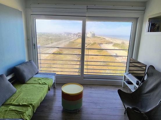 Auf dem Bild ist ein Raum zu sehen, dessen Vorderseite komplett aus einem großen Fenster besteht, dass in Richtung  Strand zeigt.
Im Zimmer selbst gibt es an der linken Wand nur ein einfaches graues Sofa mit einem grünen Überwurf davor ein kleines rundes Tischchen, an der anderen Wand stehen zwei graue hohe Campingsessel und ein weißes Tischchen, mit einem Fernseher darauf.