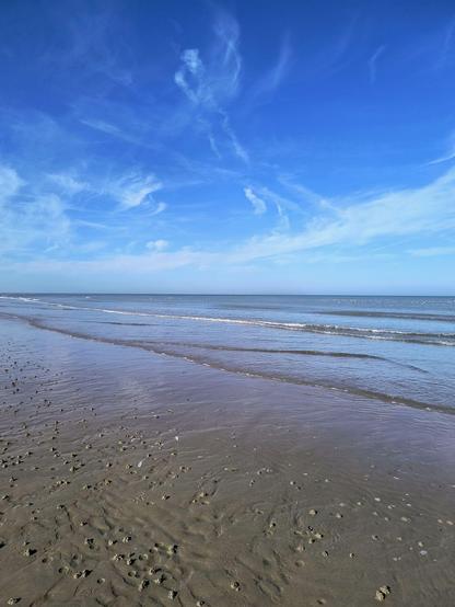 Zu sehen ist der nasse Strand und das türkisblaue Wasser und ein strahlend blauer Himmel mit wenigen zarten weißen Schleierwolken.