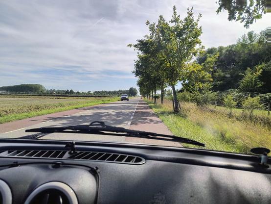 Auf dem Bild zu sehen ist der Blick aus der Frontscheibe des Aixam: auf einer schmalen Landstraße, die links und rechts einen rosaroten Fahrradstreifen besitzt, den der Aixam jetzt befahren muss, da von vorne ein PKW kommt, de auch ausweichen muss
