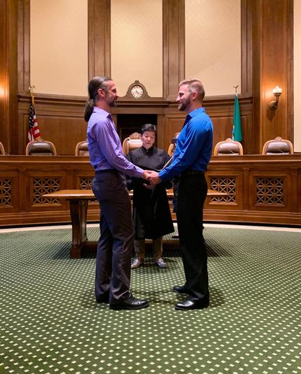 Two men stand hand in hand facing each other (Daniel in a purple shirt on the left, Casey in a blue shirt on the right) in front of Justice Yu in the Temple of Justice.