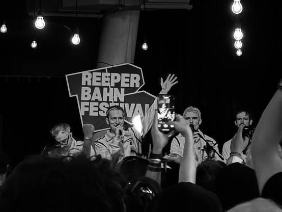 s/w Aufnahme von Kraftklub auf der Bühne vom Mojo Jazz Café. Im Hintergrund ein Reeperbahn Festival Logo, davor dicht gedrängtes Publikum.