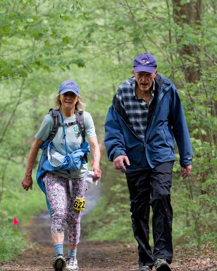 Older man and women look like a couple running in the woods.  Both are wearing billed caps.  On the left a woman wearing paisley leggings and light blue socks and shirt with a darker blue jacket wrapper around her waist.  On the right a man  is a bit huncyed over and has dark pants, a plaid shirt and a darker blue jacket than the woman.