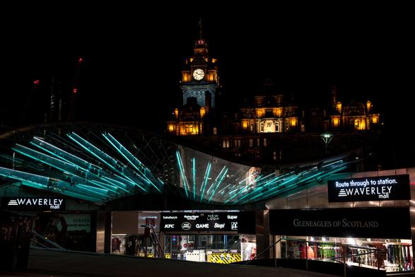 In der Dunkelheit der Nacht wird eine beeindruckende Gebäudekulisse sichtbar. Im Zentrum steht ein grosser, traditioneller Uhrturm mit einer leuchtenden Uhr. Die Fassade des Gebäudes ist hell beleuchtet und zeigt zahlreiche Fenster sowie architektonische Details. Davor erstreckt sich eine moderne, gewölbte Glasüberdachung. Diese ist von innen heraus mit strahlend türkisblauen Lichtbändern durchzogen, die wie Sonnenstrahlen wirken. Unter der Überdachung sind die Eingänge zu einem Einkaufszentrum zu erkennen, erkennbar an mehreren beleuchteten Schildern mit Namen wie "WAVERLEY mall", Oasis, KFC, GAME und New Look. Im Vordergrund sind auch Schilder für Gleneagles of Scotland und Sainsbury's angebracht. Rechts unten weist ein weiteres beleuchtetes Schild den "Route through to station" (Weg zum Bahnhof) aus.