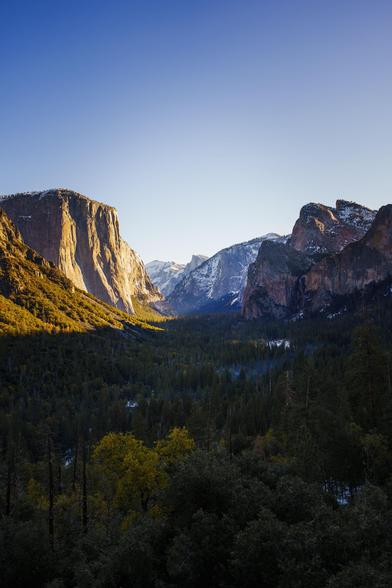 A panoramic view of a valley surrounded by towering mountains, bathed in warm sunlight under a clear blue sky.