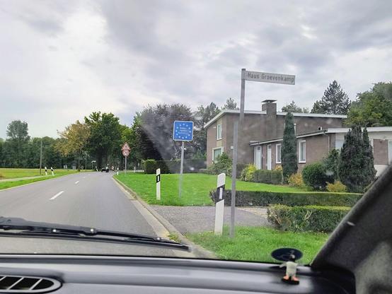 Auf dem Bild ist der Blick aus dem #Aixam zu sehen, wie eine Landstraße passiert wird. Auf der rechten Straßenseite steht ein Schild, dass anzeigt, dass die Grenze zur Bundesrepublik Deutschland passiert wurde.