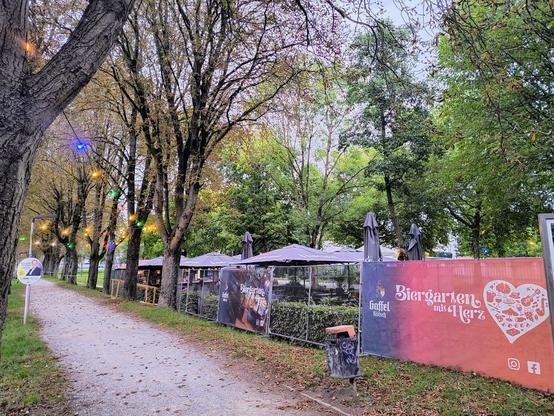 Auf dem Bild ist ein Waldweg zu sehen, an deren rechter Seite ein Biergarten zu sehen ist. Auf einem Transparent vor dem Eingang ist zu lesen " Biergarten mit Herz 
In den Bäumen neben dem Biergarten hängen bunte Lichter