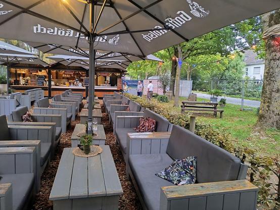 Auf dem Bild zu sehen sind Holzsofas, mit grauen Sitz und Rückenkissen in einem Biergarten, die in einer langen Reihe nebeneinander und auch jeweils gegenüber stehen, dazwischen mit Pflanzen dekorierte Tische.
Über den Sitzgruppen sind rechteckige  Sonnenschirme mit Bierwerbung zu sehen