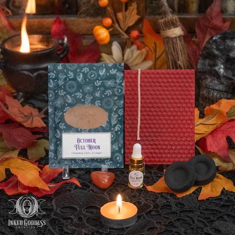 The contents of our exclusive "Aries Full Moon Ritual Kit for October 7th" product are arranged on a lace-covered surface and surrounded by faux fall foliage. They include a sheet of red beeswax and a wick; a pouch of October Full Moon herbs, including cinnamon, clove, and ginger; a dropper vial of our exclusive Full Moon Spell Oil; a tumbled carnelian gemstone; and two charcoal discs. Presented by Inked Goddess Creations.