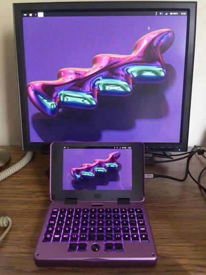 A small purple laptop sitting on a desk in front of a computer monitor.