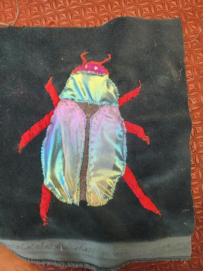 An applique of a glossy, irridescent beetle on a dark green velvet background. 