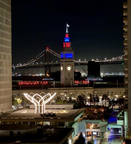 View to the east of the San Francisco ferry building at night. The clock tower is lit up red and blue (apparently something to do with the Laver Cup and the Bay Bridge is lit up in the background.