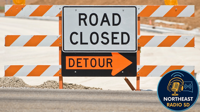 Road closed sign with orange and white stripes and a detour arrow on a construction site. "Northeast Radio SD" logo on bottom right.