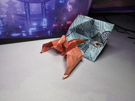 The image features two origami creations placed on a light gray surface. In the foreground, there is an orange origami figure with pointed limbs, resembling a drake/dragon with a dynamic pose. Behind it, a blue origami piece with a wave pattern and a dragon motif is visible. The background includes a screen displaying a digital image of a cityscape with traditional Asian architecture and neon signs, adding a contrasting modern element to the scene. The origami figures are intricately folded, showcasing the art of paper folding. The overall composition highlights the craftsmanship of the origami pieces against a backdrop that blends traditional and modern elements