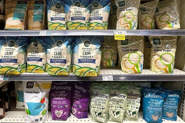 Bags of organic cane sugar on store shelves, featuring brands like 365 and Wholesome. Labels show "Regenerative" and "Fair Trade."