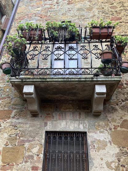 An ornate balcony with multiple potted plants rests above a wooden window, set against a textured stone wall. The balcony features intricate black ironwork, adding charm to the rustic exterior.