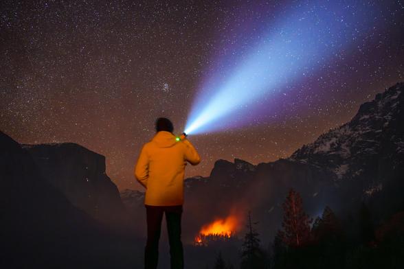 A person in a yellow jacket uses a flashlight to illuminate the starry night sky, with mountains and a distant fire visible.