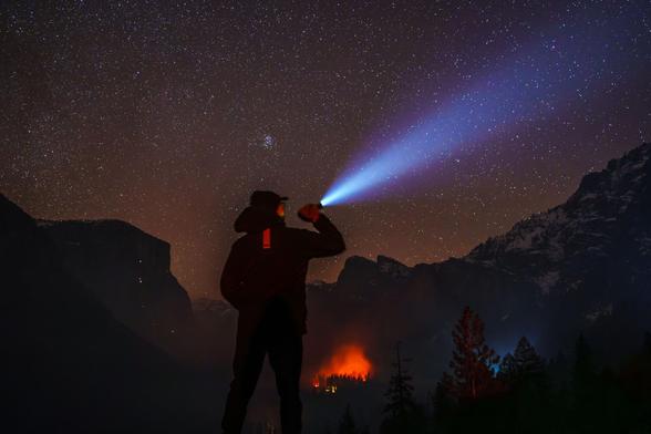 A person in a dark jacket shines a flashlight into the night sky, surrounded by mountains and a distant glowing fire.