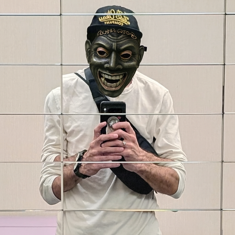 A mirror selfie segmented by horizontal tile lines shows a person wearing a black cap that reads “The Old Used Company,” a cream-colored long-sleeve shirt, and a black crossbody bag. Their face is overlaid with a dark green demonic mask, resembling a theatrical or tribal design with exaggerated features and gold detailing. The mask adds a surreal, performative layer to the otherwise casual, reflective image.