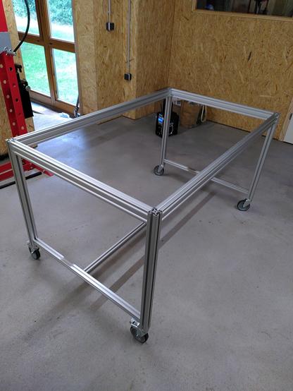 Silver aluminium table frame with wheels photographed in a work shop with a grey concrete floor and wooden walls.