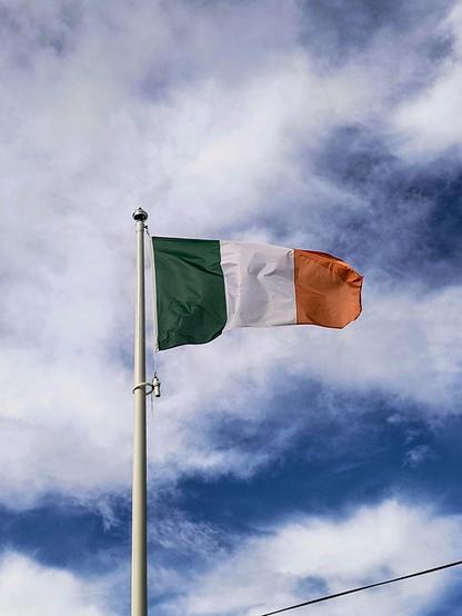 Zu sehen ist die Flagge von Irland an einem Fahnenmast, wie sie bei leicht bewölktem blauen Himmel im Wind weht