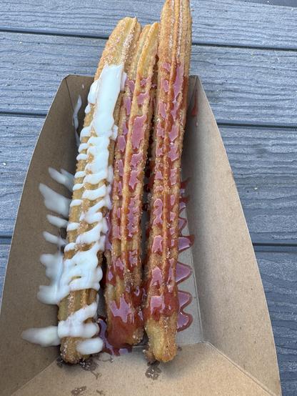 Three churros in a cardboard container. Two have strawberry sauce on them and one has vanilla sauce drizzled on it. They were both good, but the vanilla was a little better!