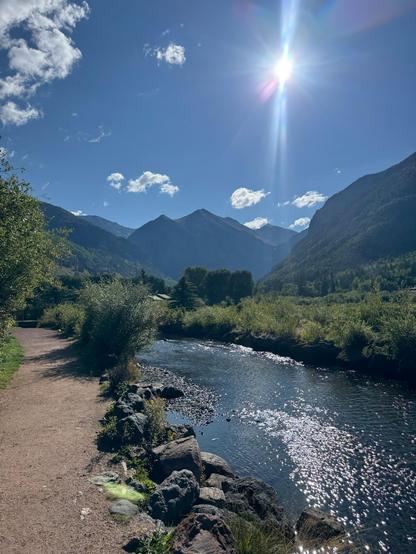 Photo of a narrow river, running past a trail in western Colorado. The sun is low in the autumn afternoon, sky, and glinting off the slowly moving water of the river. There are a few small puffs of clouds in an otherwise bright blue sky. 