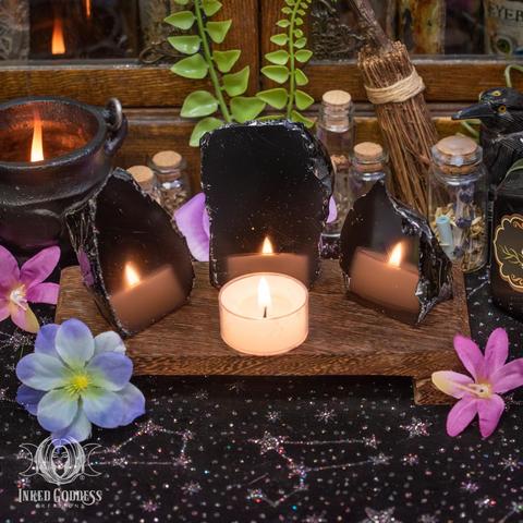 Three specimens of the "Obsidian Standing Scrying Stone" product are arranged on a wooden platform facing a tealight candle. They are flat and highly polished on one side, while the rest of the stone is in its natural jagged state. Presented by Inked Goddess Creations.