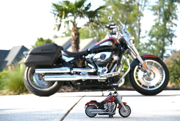A small LEGO version of a Harley-Davidson motorcycle on a white driveway. Behind it, slightly out of focus is the actual version of the same motorcycle. There are green trees and houses in the background.