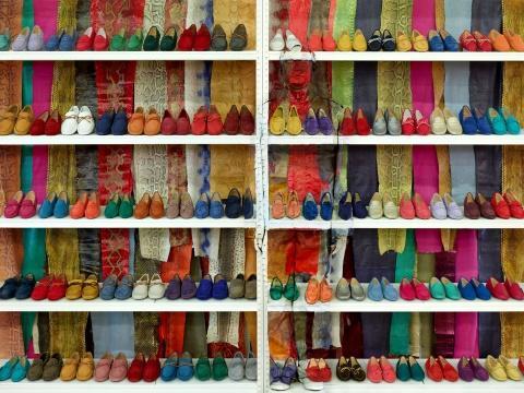 Liu Bolin stands camouflaged in front of white shelving stacked with colorful shoes and scarves. His body is painted to match the background, making him nearly invisible—only faint shadows and outlines reveal his presence.