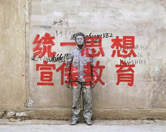 Liu Bolin stands in front of a concrete wall with large red Chinese characters promoting state ideology. His face and body are painted to blend perfectly with the text and surface, rendering him nearly invisible except for subtle outlines.