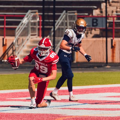 A football player wearing red gear with the number 89 has his left (your right) knee on the ground and his other leg bent with his left hand on it and a football held in his right gloved hand in the endzone while a player in a white jersey, blue pants and brown helmet stands behind him,  past a green patch note some empty bleachers,  a stair up,  and the letter B denoting the section