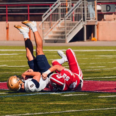 By section D of the stands, a football player wearing red pants, jersey and helmet and the number 52 grabs tight onto number 9 of the opposing team wearing a beige helmet, white jersey and blue pants who grips the ball tightly as his feet point straight up in the air