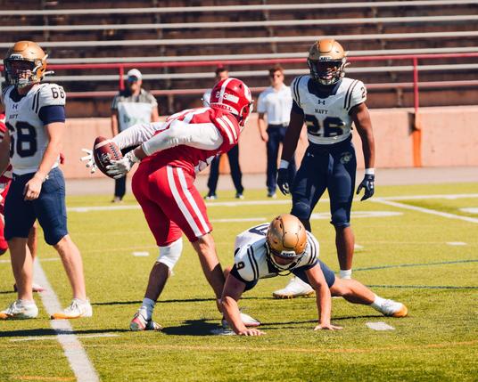 A player wearing red gear with a white undershirt holds a football behind himself that he snatched away from the player right in front of him who is bent forward trying to snatch it himself while number 28 from Navy and three people on the sideline look on.