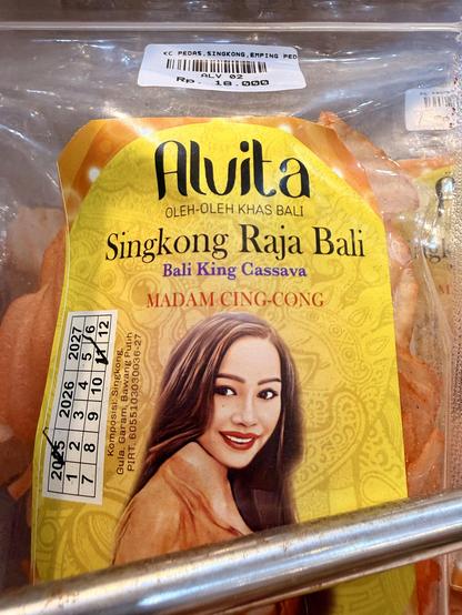 Foto einer Snackverpackung aus Bali. Auf der Vorderseite steht in großen Buchstaben ‚Singkong Raja Bali – Bali King Cassava‘. Darunter der Markenname ‚Alvita‘ und der Schriftzug ‚Madam Cing-Cong‘. Abgebildet ist das Porträt einer lächelnden Frau mit langen, dunklen Haaren. In der durchsichtigen Verpackung erkennt man dünne, orangefarbene Chips aus Maniok (Cassava).