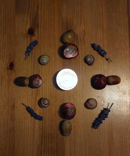 A lit tea light on a wooden table, surrounded by 4 each of conkers, old acorns, snail shells, and sprigs of lavender arranged around the tea light.