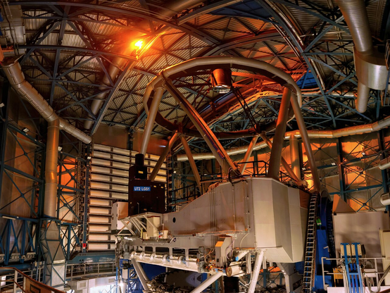 The image shows the interior of a dome with strut-reinforced walls. Centred bit shifted to the right there is a bulky object, a telescope, with a round metallic structure on the top. To the left of this is a black rectangular block with the letters “UT2 LGS 1”, projecting an orange beam that reaches the top of the dome.