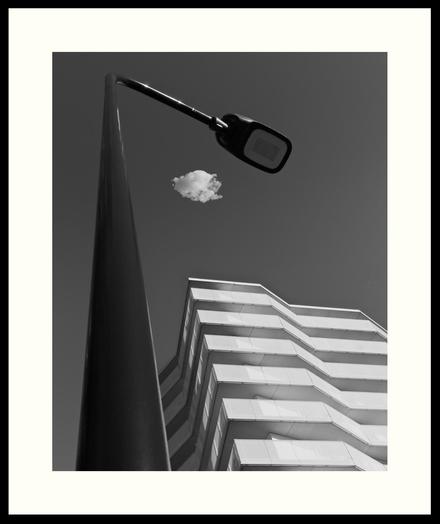 b&w - A simple, three object image, that shows a bright white multi story residential building, which is almost dwarfed by a dark streetlamp. A single cloud is caught between the the building and the streetlamp.